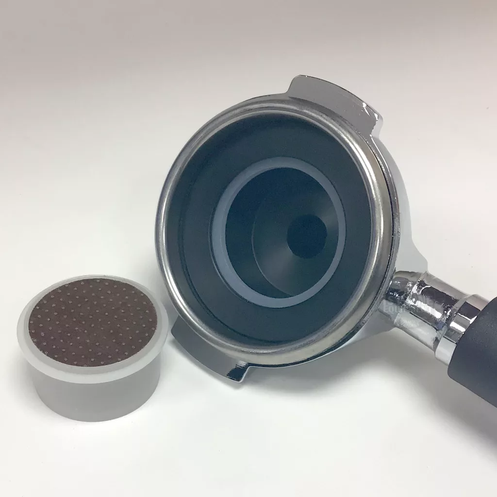 A Lelit PLA583S 58mm Pressurized Espresso Point Capsule Portafilter with a black handle rests next to a round, perforated coffee puck on a white background. © 2025 1st-line.com
