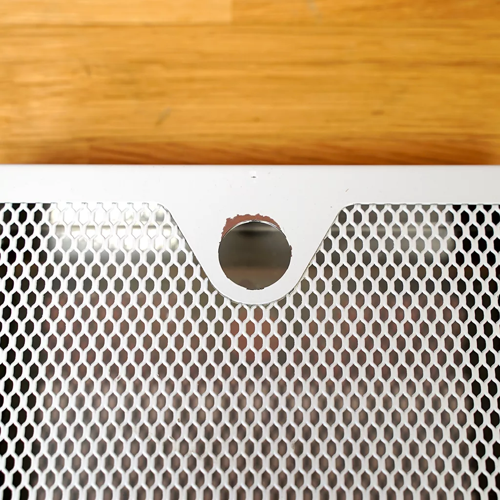 A close-up of the SIDEWALK SALE-VBM White Junior WHITE Drip Tray, Grill & Water Tank Cover E209, featuring a white metal mesh with a round notch and chipped paint, set against a wooden background. © 2025 1st-line.com