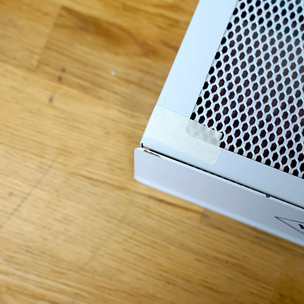 A SIDEWALK SALE-VBM White Junior WHITE Drip Tray, Grill & Water Tank Cover E209 sits on a light wooden floor. A piece of white tape is affixed to one corner of the metal cover, which features a perforated vent design. © 2025 1st-line.com