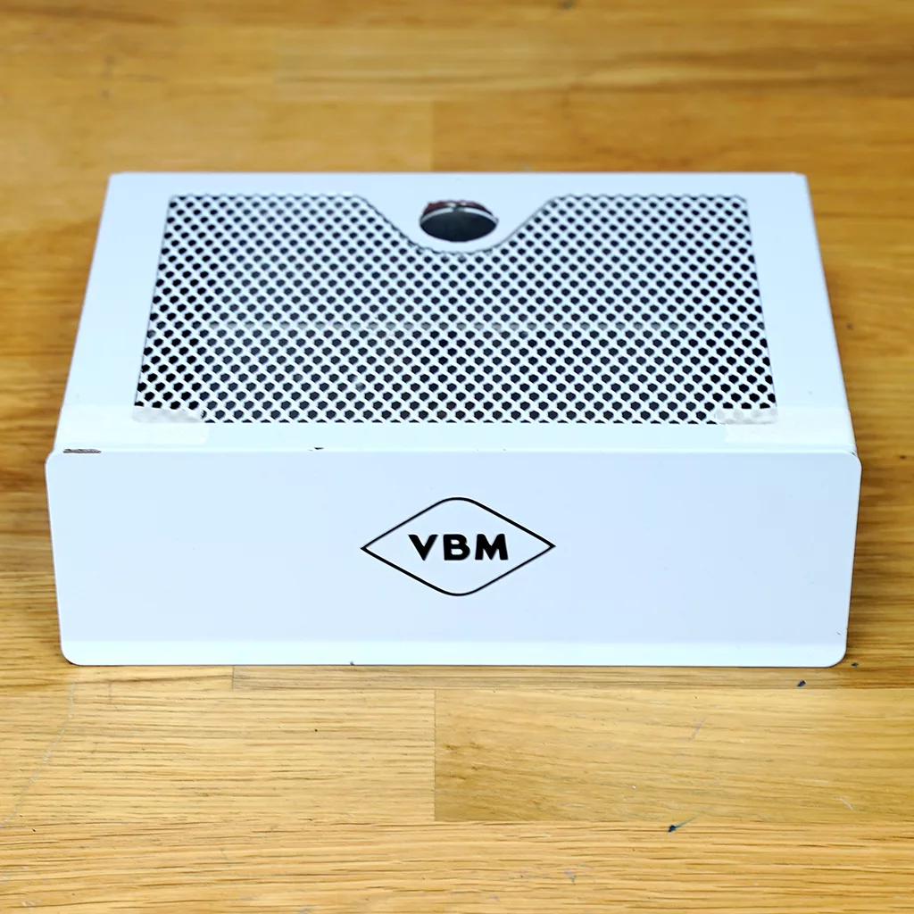 A SIDEWALK SALE-VBM White Junior WHITE Drip Tray, Grill & Water Tank Cover E209 sits on a wooden surface. The rectangular metal box has a perforated grill top with a central hole and features a diamond-shaped VBM logo on the front. © 2025 1st-line.com