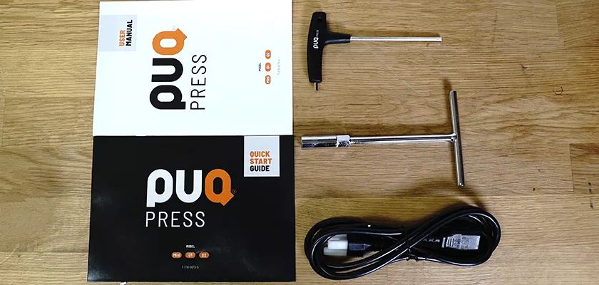 Included with the DISCONTINUED PUQPRess Mini Home Precision Coffee Tamper (53.0–58.3mm) are a user manual, quick start guide, T-handle hex key, socket wrench, and power cable—all displayed on a wooden surface. © 2025 1st-line.com