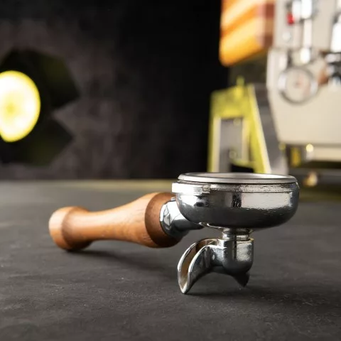 A metal espresso portafilter with a wooden handle and premium accessories rests on a dark countertop, with the DISCONTINUED Elektra Verve Dual Boiler Espresso Machine and light softly blurred in the background. © 2025 1st-line.com