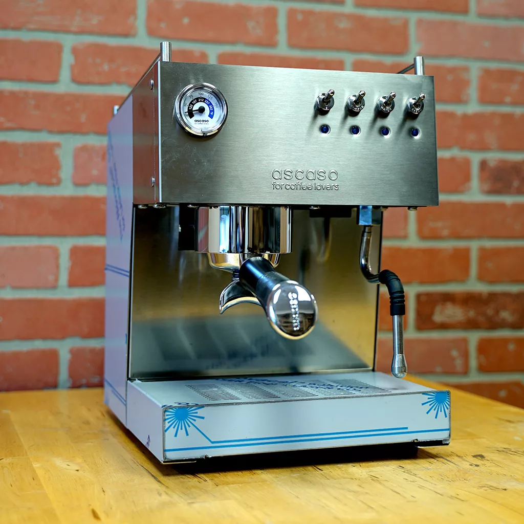 A returned and refurbished Ascaso Steel Uno Professional V2 SHINY Mirror Finish Espresso Machine is displayed on a wooden counter in front of a red brick wall. © 2025 1st-line.com