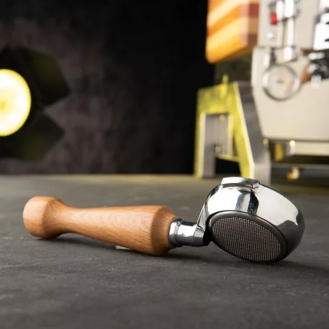 A wooden-handled portafilter rests on a dark counter beside the discontinued Elektra Verve Dual Boiler Espresso Machine with premium accessories, with a blurred yellow light glowing softly in the background. © 2025 1st-line.com
