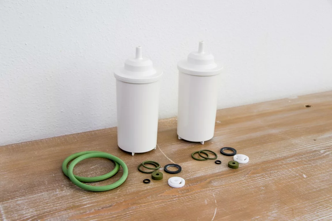 Two white cylindrical filter cartridges with top nozzles sit on wood, each paired with green and black O-rings, washers, and white plastic parts—premium accessories from the discontinued Elektra Verve Dual Boiler Espresso Machine. © 2025 1st-line.com