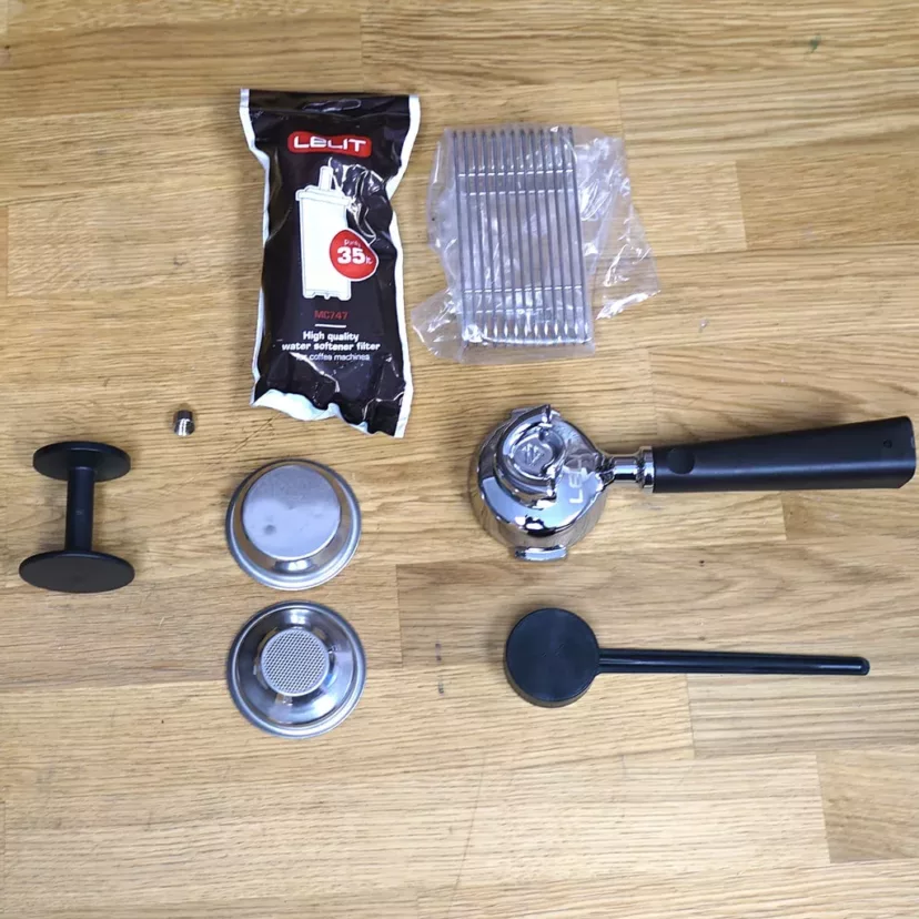 A selection of espresso accessories on a wooden surface—ideal for the (SOLD) SIDEWALK SALE- Lelit PL82T Kate Espresso Machine with Built-in Grinder (returned & refurbished): portafilter, tamper, filter baskets, cleaning brush/powder, and metal tool. © 2025 1st-line.com
