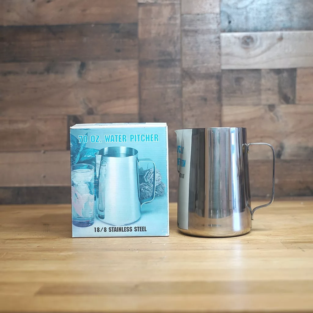 A SIDEWALK SALE - Water Pitcher 70 OZ. sits on a wooden surface beside its box, which displays an image and product specs. A wood-paneled wall is visible in the background—an ideal find for your next Sidewalk Sale!. © 2025 1st-line.com