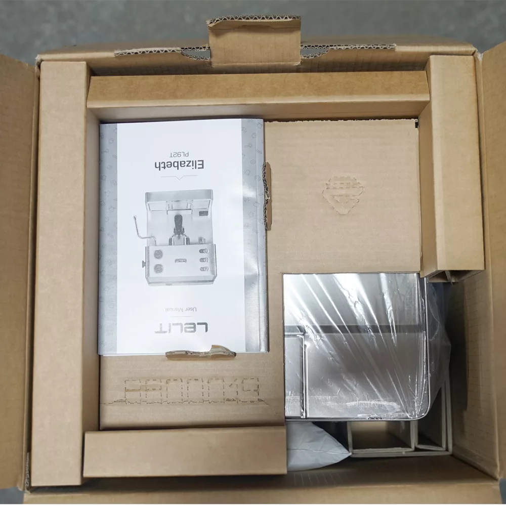 An overhead view of Lelit Elizabeth packaging shows a partially unpacked coffee machine in a cardboard box, with the user manual labeled ELIZABETH and metal components wrapped in plastic beside it. © 2025 1st-line.com