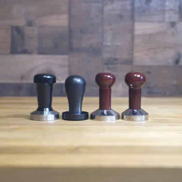 Four SIDEWALK SALE - Tampers—two with black handles and two with dark brown—are neatly arranged on a wooden surface, making them the perfect addition to your coffee setup. © 2025 1st-line.com