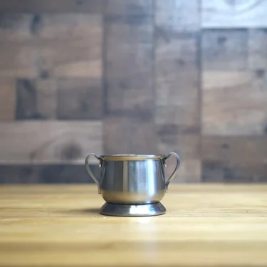 The SIDEWALK SALE - Sugar Bowl, a small silver piece with two handles, rests on a wooden surface against a blurred wood background—perfect for stylish displays. © 2025 1st-line.com