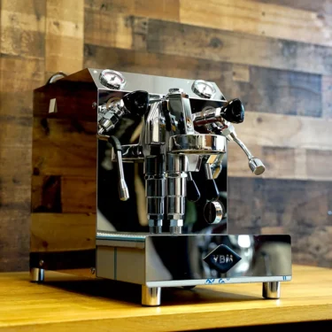 A SIDEWALK SALE - VBM Domobar Super HX Tank Espresso Machine DEMO (E114) shines on a wooden countertop, with dual gauges and steam wand, set against a rustic wall and ready to impress at your next sidewalk sale. © 2025 1st-line.com