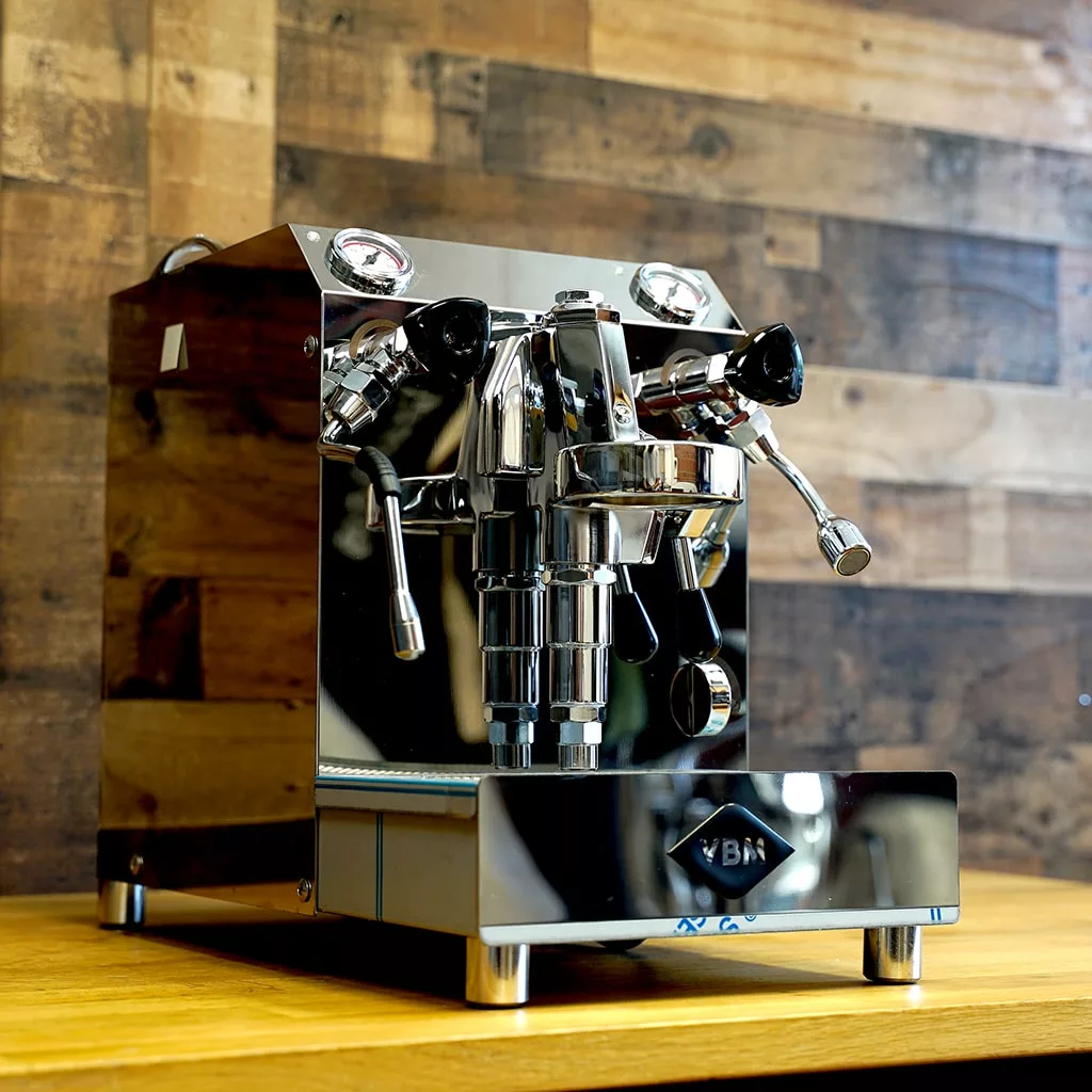 A SIDEWALK SALE - VBM Domobar Super HX Tank Espresso Machine DEMO (E114) shines on a wooden countertop, with dual gauges and steam wand, set against a rustic wall and ready to impress at your next sidewalk sale. © 2025 1st-line.com