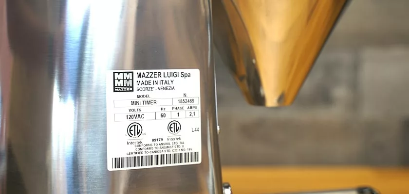 Close-up of a Mazzer Mini Timer Converted-to-Doserless Espresso Coffee Grinder label, showing model info and certifications, with the metallic doserless grinder and hopper from the Mazzer Mini line in the background. © 2025 1st-line.com