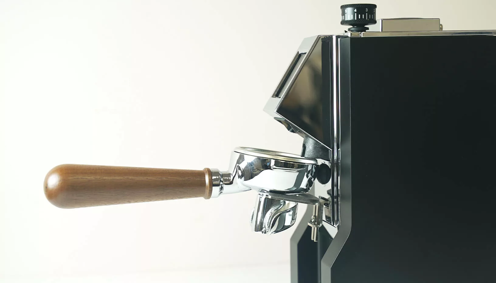 A close-up side view of a modern espresso machine with a wooden-handled portafilter, set against a plain light background, next to the Eureka Oro Mignon XL Espresso Coffee Grinder. © 2025 1st-line.com