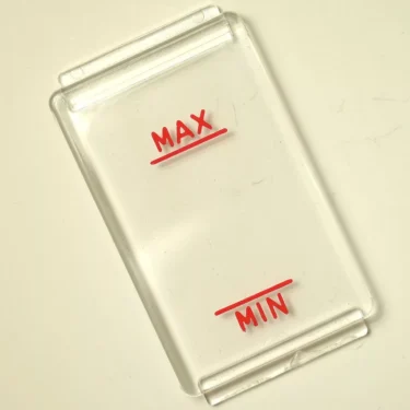 A clear rectangular plastic Rancilio Level Mask 90x50 mm, featuring red underlined MAX and MIN markings, is shown on a light surface, likely serving as a gauge or indicator cover. © 2025 1st-line.com