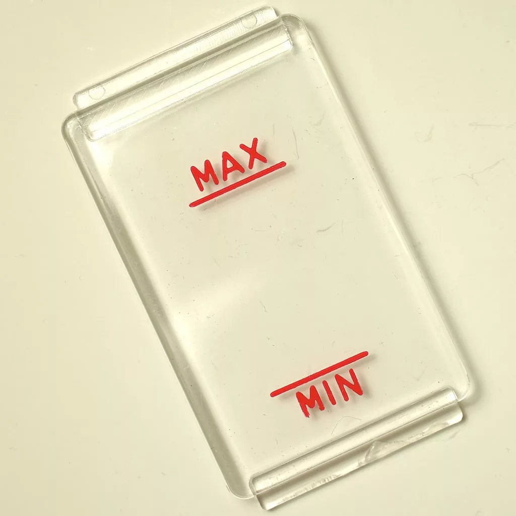 A clear rectangular plastic Rancilio Level Mask 90x50 mm, featuring red underlined MAX and MIN markings, is shown on a light surface, likely serving as a gauge or indicator cover. © 2025 1st-line.com