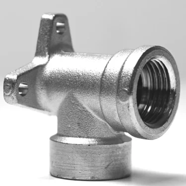 A close-up of the V.1872 1/2Mx1/2F Elbow Fitting shows its metallic finish, threaded opening, and two mounting holes on a flat extension, all set against a plain background. © 2025 1st-line.com