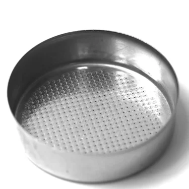 A round metal sieve with fine mesh holes sits on a white surface, its shiny rim reflecting light. Next to it is the P..455 GASKET 15x6x6mm, placed neatly beside the clean and empty sieve. © 2025 1st-line.com