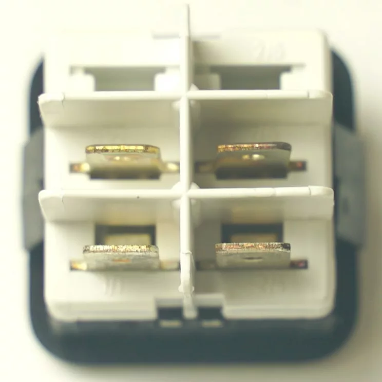 Close-up of a V.4401 MOMENTARY ROCKER SWITCH - CLEAR, featuring a white and black electrical connector with four metal prongs arranged in a square, separated by plastic dividers. © 2025 1st-line.com