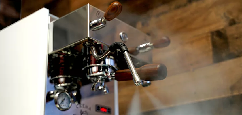 A close-up of a shiny Bezzera Crema PM HX Espresso Machine with wooden handles, pressure gauge, and steam from the metal steam wand, set against a wooden wall background. © 2025 1st-line.com