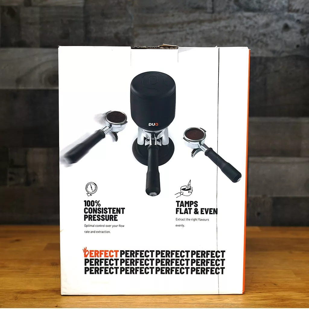 A product box on a wooden table displays the *SOLD* - SIDEWALK SALE - PUQPress Mini Home Precision Coffee Tamper - 58.0mm OPEN BOX E113, with two portafilters. Text highlights 100% consistent pressure, tamps flat & even, and repeats Perfect. © 2025 1st-line.com