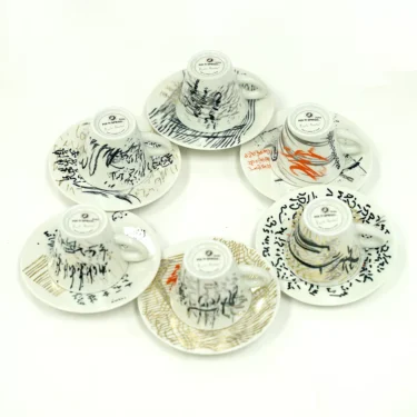 Six white teacups from the Francis!Francis! Encrypted Collector Espresso Set, each with abstract black, orange, and gold patterns, sit upside-down on saucers in a loose circle on a plain white background. © 2025 1st-line.com