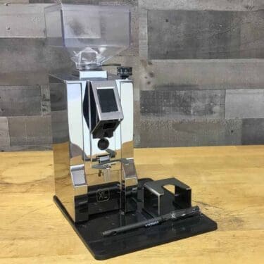 A SIDEWALK SALE Eureka Oro Mignon XL On Demand Espresso Coffee Grinder in chrome (OPEN BOX E209) sits on a wooden counter with a detachable black tray and accessories in front, backed by a gray wooden wall. © 2025 1st-line.com