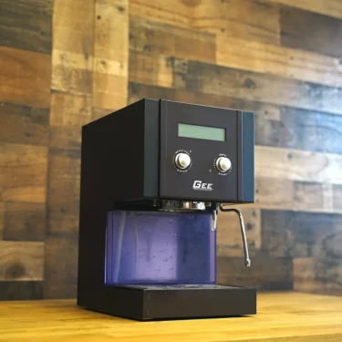 A SIDEWALK SALE - GEE Digital Espresso Coffee Machine PARTS ONLY (E213), sleek and black with a digital display and two knobs, sits on a wooden surface against a wood-textured wall. © 2025 1st-line.com