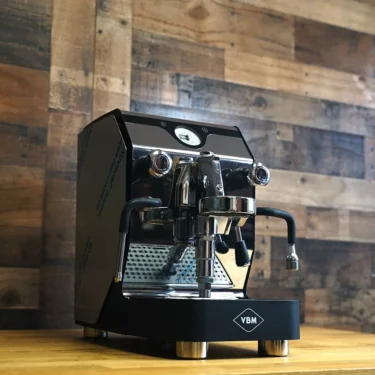 An open box SIDEWALK SALE - VBM Domobar Super Analogic HX Espresso Machine (E013) sits on a wooden countertop against a rustic wall, featuring dual spouts, pressure gauge, and metal steam and hot water wands. © 2025 1st-line.com