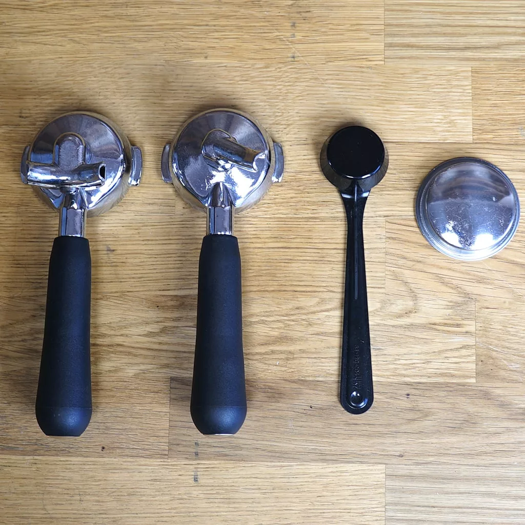 Two portafilters with black handles, a black tamper, and a metal filter basket sit on a wooden surface—ideal accessories for your SIDEWALK SALE - VBM Domobar Super Analogic HX Espresso Machine OPEN BOX (E013). © 2025 1st-line.com