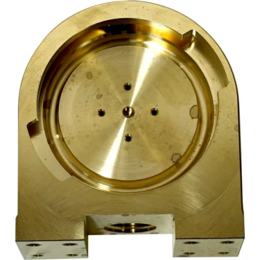 A close-up of the Elektra 05906014 Delivery Group Body, a machined gold metal part featuring a circular recessed center, small holes, and a flat base with four screw holes. © 2025 1st-line.com