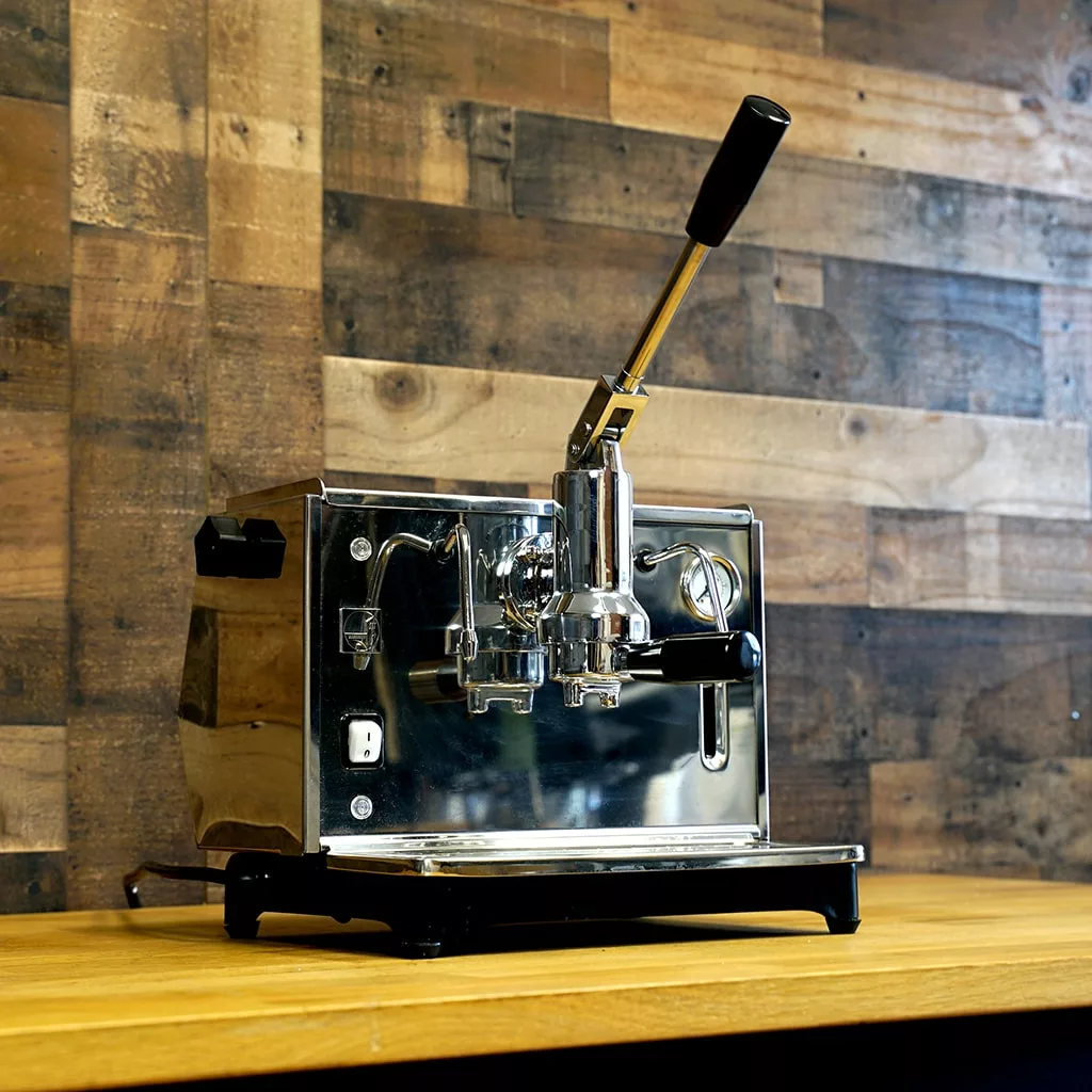 A refurbished Ponte Vecchio Lusso 1 Group Spring Piston Lever Espresso Machine (SOLD) sits on a wooden countertop, its black handle contrasting with the wood-paneled wall background. © 2025 1st-line.com