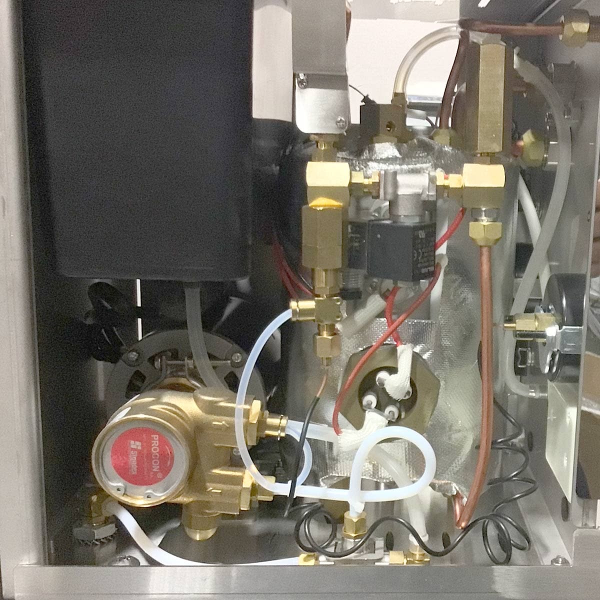 Close-up of internal components in the Bellezza Inizio R Leva HX Espresso Machine, with brass valves, a pump, wires, tubes, and connectors neatly arranged within its metal enclosure. © 2025 1st-line.com