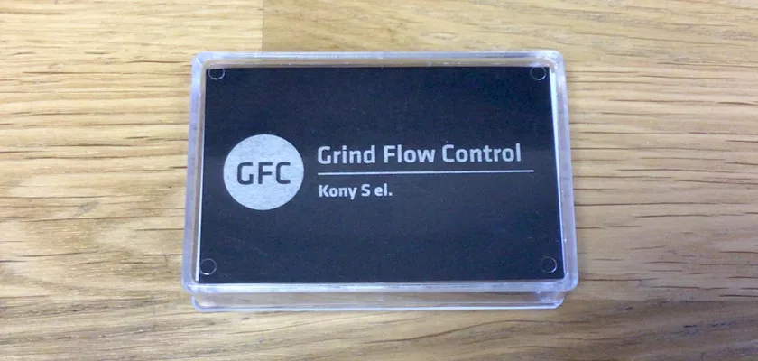 A black rectangular plaque with GFC Grind Flow Control Kony S.el. in white, encased in clear plastic, rests on a light wooden surface—ideal for the Mazzer Kony S Time-Based Commercial Espresso Coffee Grinder. © 2025 1st-line.com
