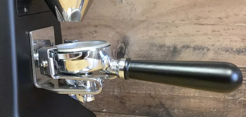 A close-up of a portafilter attached to an espresso machine, with a Mazzer Kony S Time-Based Commercial Espresso Coffee Grinder blurred in the background and a wooden surface behind. © 2025 1st-line.com