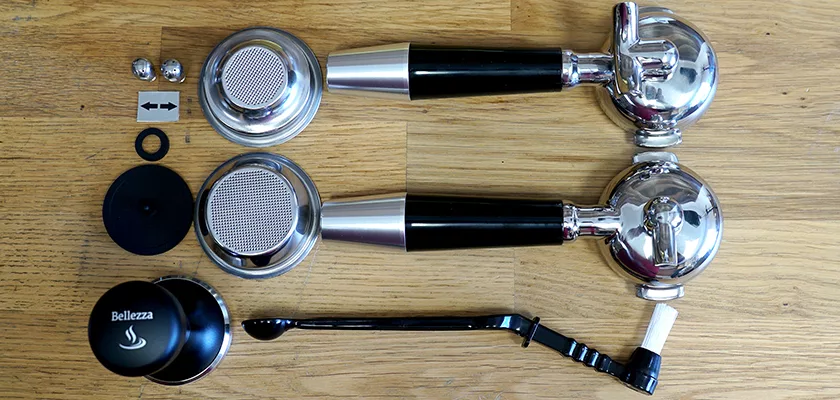 Accessories for the REDIRECTED Bellezza Inizio R Leva HX Espresso Machine—including portafilters, filter baskets, tamper, dosing spoon, gaskets, a cleaning brush, and metal screws—are arranged neatly on a wooden surface. © 2025 1st-line.com