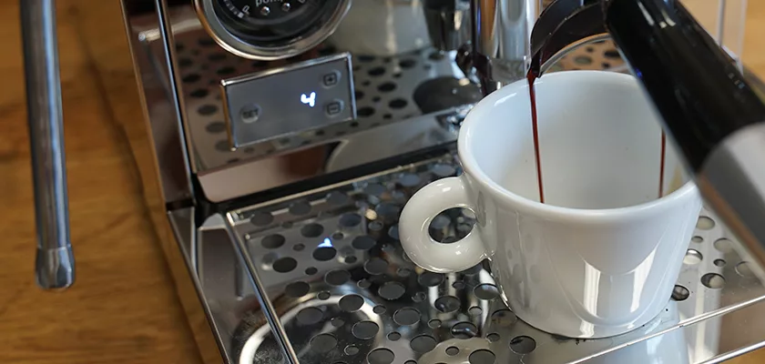 Close-up of a white cup being filled with espresso from the REDIRECTED Bellezza Inizio R Leva HX Espresso Machine, as rich coffee pours into the cup on its perforated metal drip tray. © 2025 1st-line.com