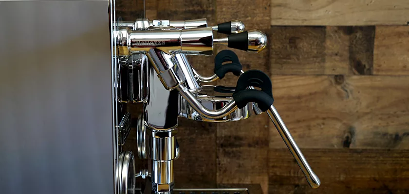 Close-up of the polished chrome REDIRECTED Bellezza Inizio R Leva HX Espresso Machine lever group against a wooden wall, highlighting the Leva HX handle and mechanical components on the left. © 2025 1st-line.com