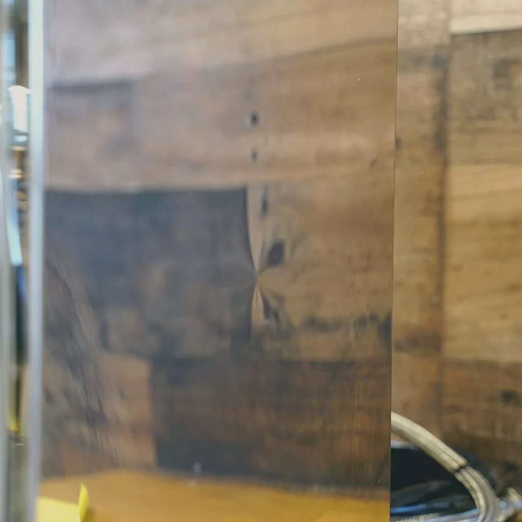 A close-up of a reflective surface before a wooden wall with visible grain, some blurred items, and part of the SIDEWALK SALE - Fiorenzato Volante Direct Connect HX Espresso Machine (E214) at the bottom edge. © 2025 1st-line.com