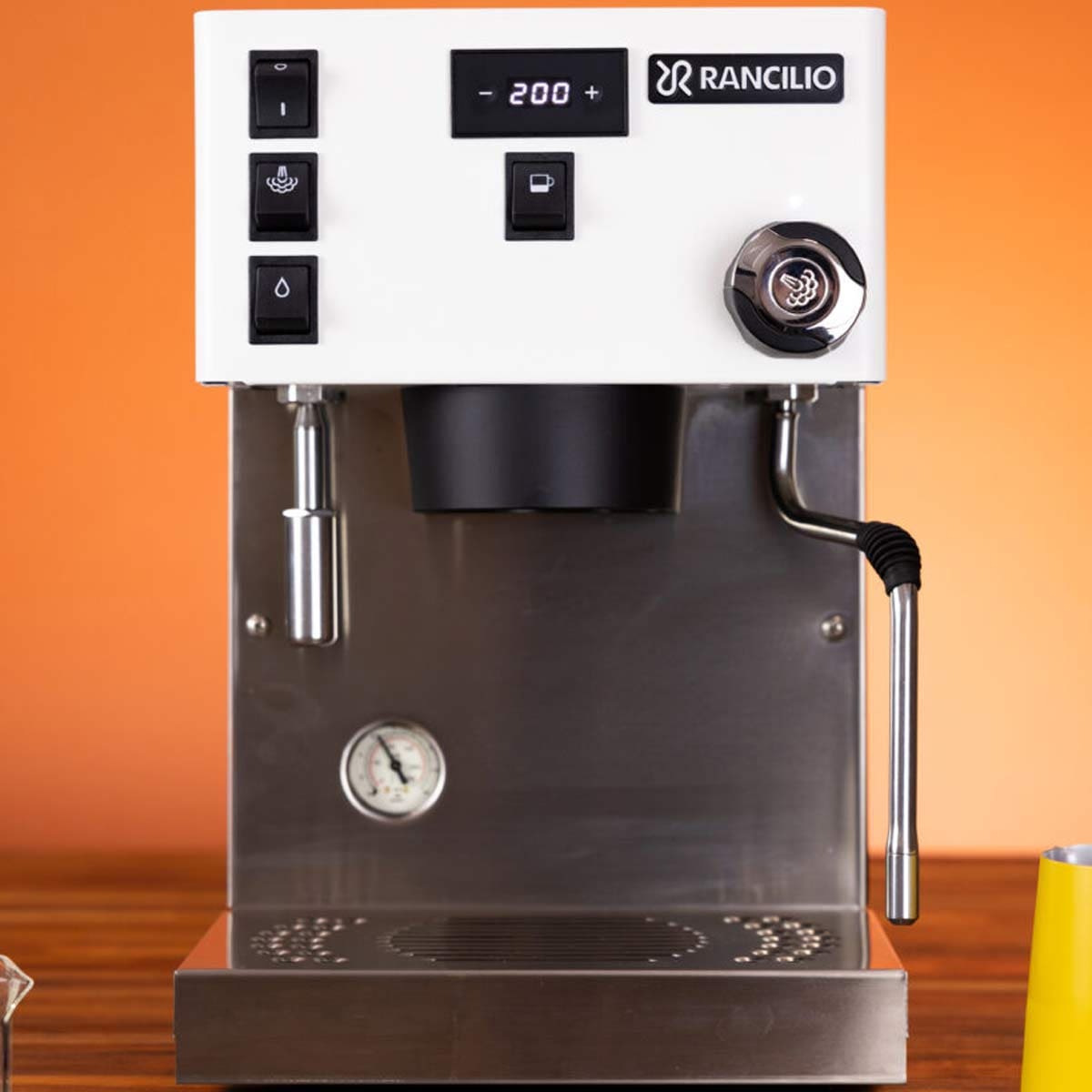 A Rancilio Silvia Pro X Dual Boiler Espresso Machine with black buttons, a pressure gauge, steam wand, and digital display sits on a wooden counter against an orange background. © 2025 1st-line.com