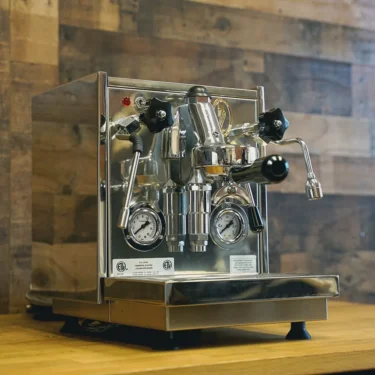 A SIDEWALK SALE - Fiorenzato Volante Direct Connect HX Espresso Machine (E214) with gauges, knobs, and levers sits on a wooden countertop in front of a rustic wood-paneled wall. © 2025 1st-line.com