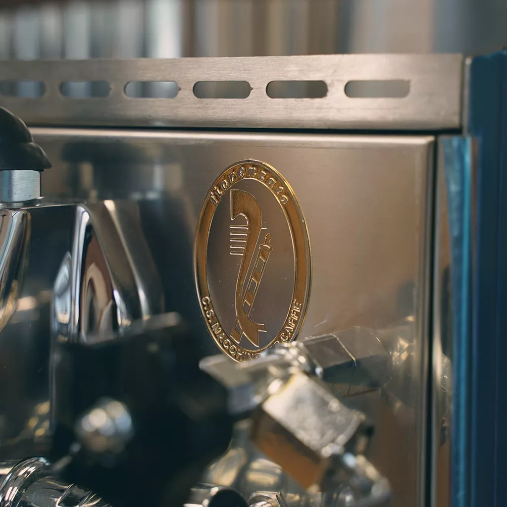 Close-up of a shiny SIDEWALK SALE - Fiorenzato Bricoletta Direct Connect HX Espresso Machine (E212) with a gold emblem featuring a coffee tamper, laurel wreath, and “TOP CRAFTSMAN COMMERCIAL QUALITY” engraved—ideal for display. © 2025 1st-line.com