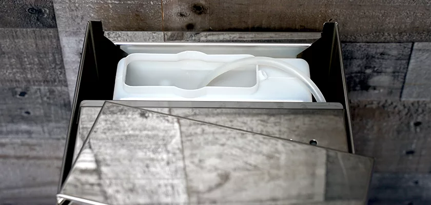 A close-up of an open metal dispenser, echoing the precision of the Bezzera Aria HX Special Edition Espresso Machine w/ Flow Control, shows a white plastic refill container with a tube against a wood-textured wall. © 2025 1st-line.com