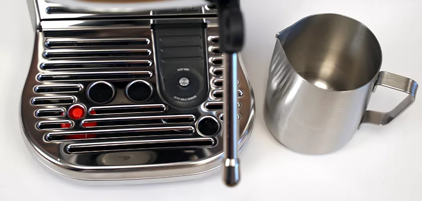 A close-up of the Breville Bambino Plus™ Espresso Machine’s stainless steel drip tray and control panel sits beside a metal milk frothing pitcher on a white surface. © 2025 1st-line.com
