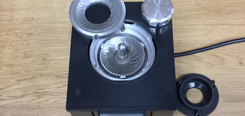 A close-up of the Macap Leo 55 Espresso Coffee Grinder, open to show its metal burrs and circular dial on a wooden surface, with a detached black ring next to it. © 2025 1st-line.com