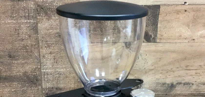 A close-up of the Macap Leo 55 Espresso Coffee Grinder shows its clear, empty hopper with a black lid on a sleek black base, set against a wooden plank background. © 2025 1st-line.com