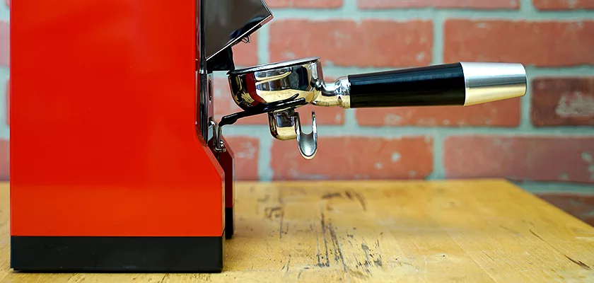 A close-up side view of a red espresso machine with a metal portafilter on a wooden surface beside an Eureka Mignon Zero Espresso Coffee Grinder, set against a brick wall background. © 2025 1st-line.com