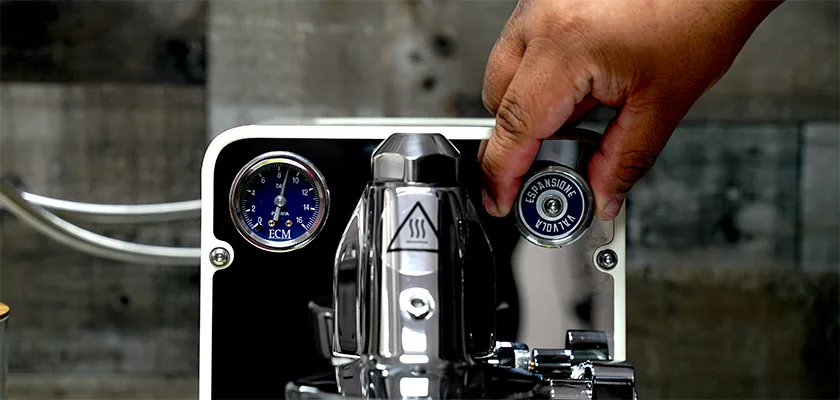 A hand turns a knob on the DISCONTINUED ECM Puristika Espresso Machine, which has a blue pressure gauge on the left and shiny stainless steel that reflects its surroundings. © 2025 1st-line.com