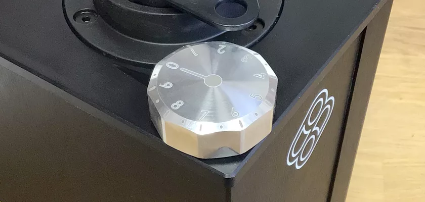 A metallic combination lock dial with engraved numbers is on the corner of a black Macap Leo 55 Espresso Coffee Grinder safe, featuring a white circular logo on the side. The background is a light wood surface. © 2025 1st-line.com