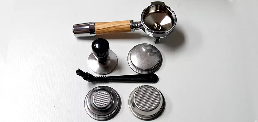 A portafilter with a wooden handle, a tamper, a cleaning brush, and four metal espresso filter baskets for the DISCONTINUED ECM Puristika Espresso Machine are arranged on a white surface. © 2025 1st-line.com